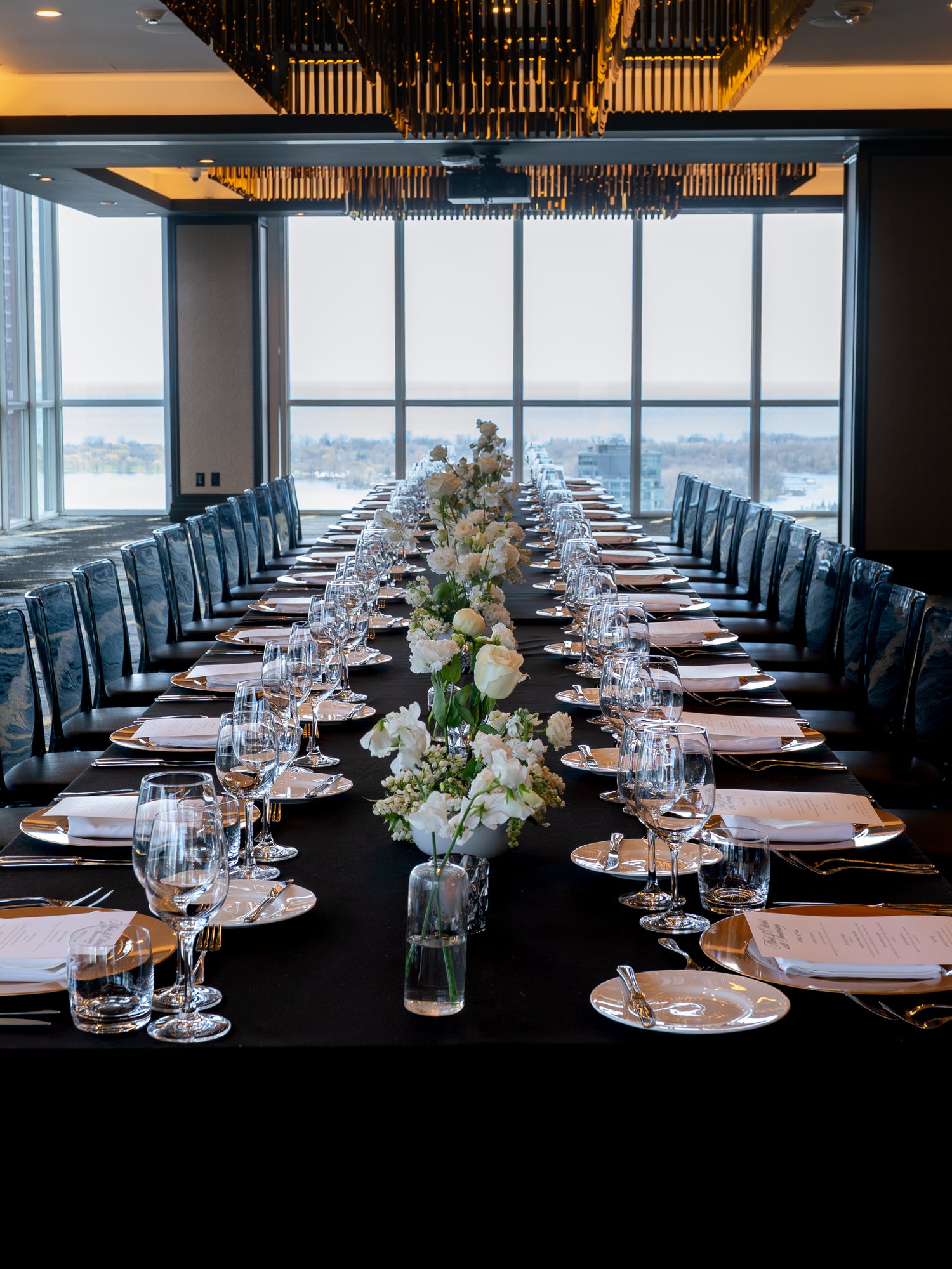 Long dining table set for a formal event with floral centerpieces, glasses, and cutlery in a room with large windows.
