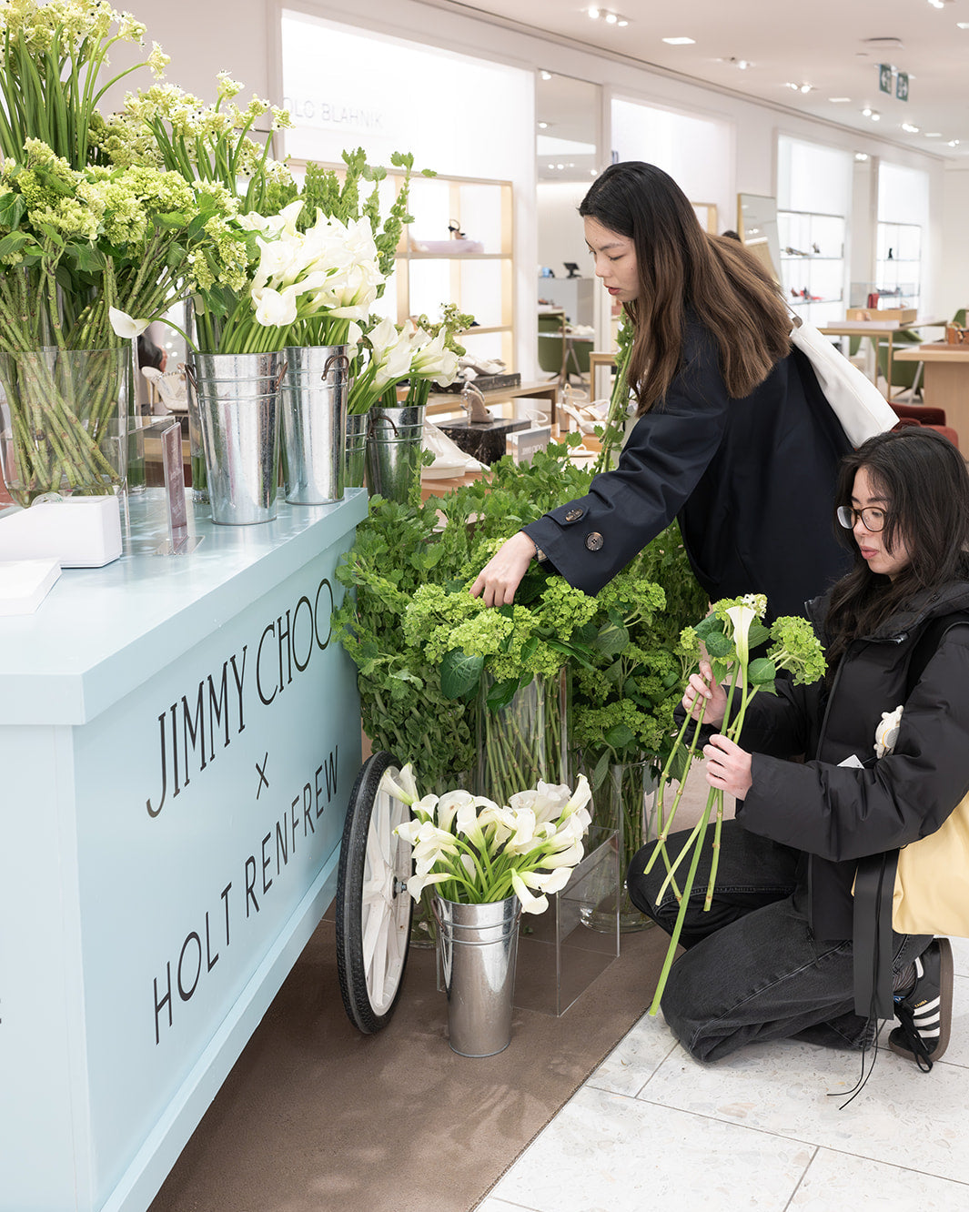 Two women arranging flowers in a store with 'Jimmy Choo x Holt Renfrew' branding.