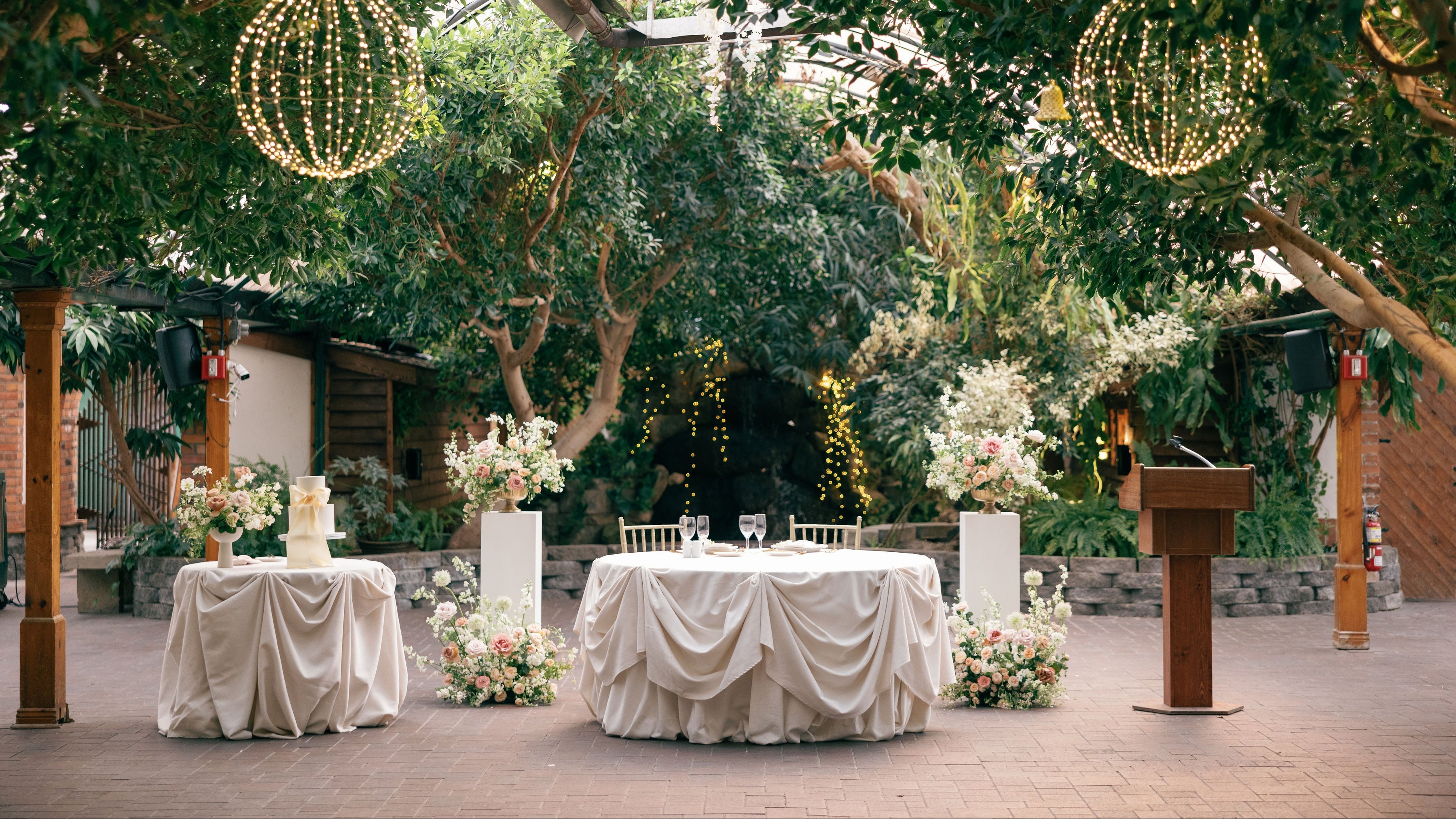 Decorated outdoor wedding setup with tables, floral arrangements, and hanging lights.